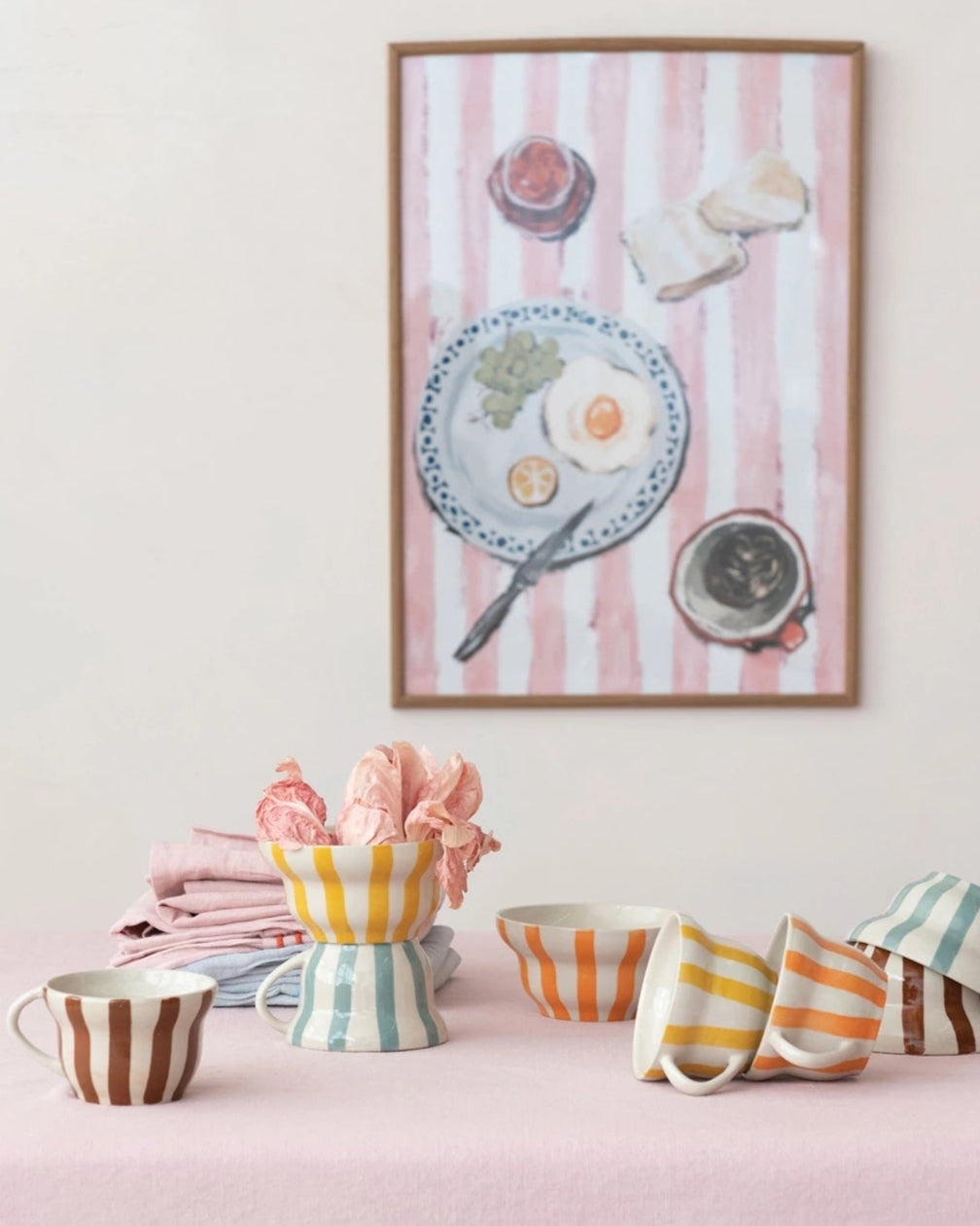 Framed artwork on a wall with striped background, below which are colorful ceramic items on a pink surface.